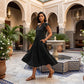 Elegant Torvun black tiered midi dress with ruffle cap sleeves and V-neck, worn by a model walking in a beautiful Moroccan-style courtyard, showcasing its flowing silhouette.