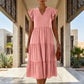 Female model walks forward in a modern architectural setting, wearing a Torvun Blush Pink tiered midi dress with V-neck and ruffle cap sleeves.