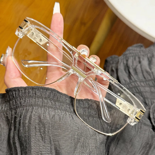 Close-up view of oversized square sunglasses with a transparent acetate frame, gold double bridge, and subtle temple accents. The clear lenses are held by a hand against a dark fabric background.