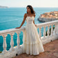 Elegant woman wears a vanilla cream textured maxi dress with ruffled neckline, spaghetti straps, bustier bodice, and a tiered skirt, posing on a balcony overlooking the blue Mediterranean Sea.