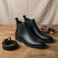 Vantor mens Chelsea boots in midnight black microfiber leather with matching belt and leather wax on a wooden table.