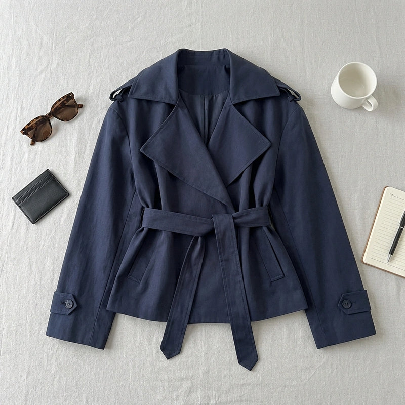 Varelle navy belted short trench coat, flat-lay view with accessories. Shows structured cotton blend fabric, wide lapels, and tie waist.