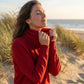 Vellura soft cowl neck sweater in red, worn by a woman on a beach — side view showing the ribbed cuffs and elegant draped neckline.