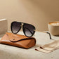 VELORA oversized aviator sunglasses with black gradient lenses and gold double bridge resting on a brown leather case next to a coffee cup.