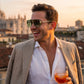 Velora retro aviator sunglasses in green acetate with gold double bridge, worn by a smiling man in a beige blazer holding a drink at sunset.