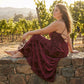 Veloura burgundy jacquard maxi dress worn in a vineyard setting, showing the floral relief texture and open-back strap detail from the side.