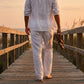Ventoso men's linen trousers in white, back view showing the relaxed straight-leg fit and breathable fabric while walking barefoot on a sunset boardwalk.