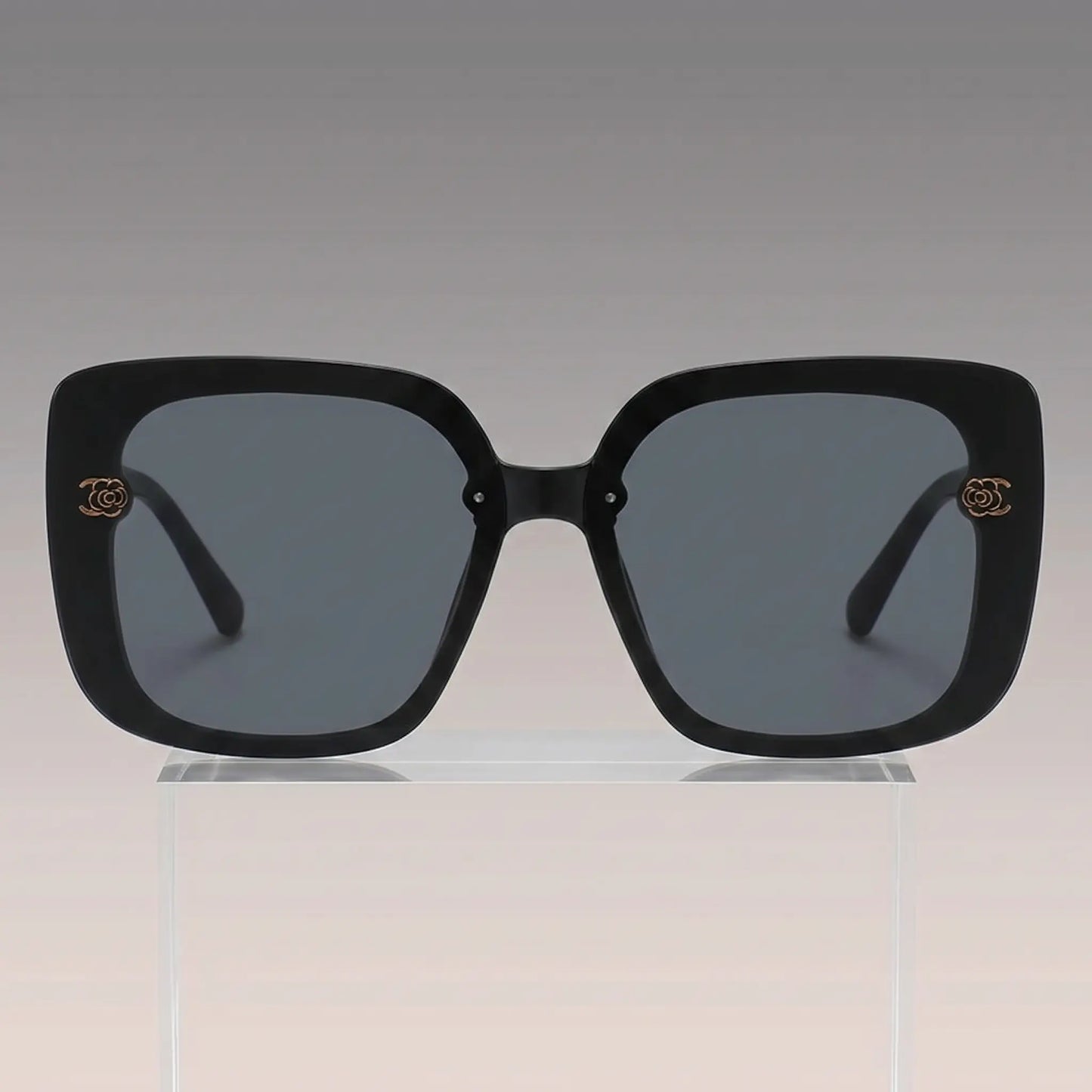 Vesryn oversized square sunglasses with thick black acetate frames and dark gradient grey lenses, featuring gold floral-inspired metal logo accents on the temples.