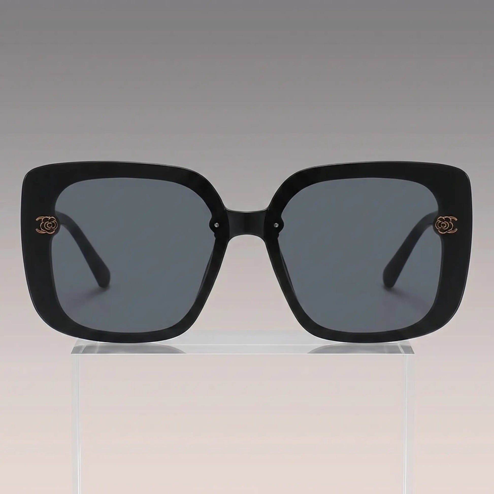 Vesryn oversized square sunglasses with thick black acetate frames and dark gradient grey lenses, featuring gold floral-inspired metal logo accents on the temples.