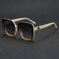 Close-up of oversized square sunglasses with off-white temples, featuring a gradient amber-brown front and gradient grey lenses, showcasing a gold floral metal logo detail on the temple.