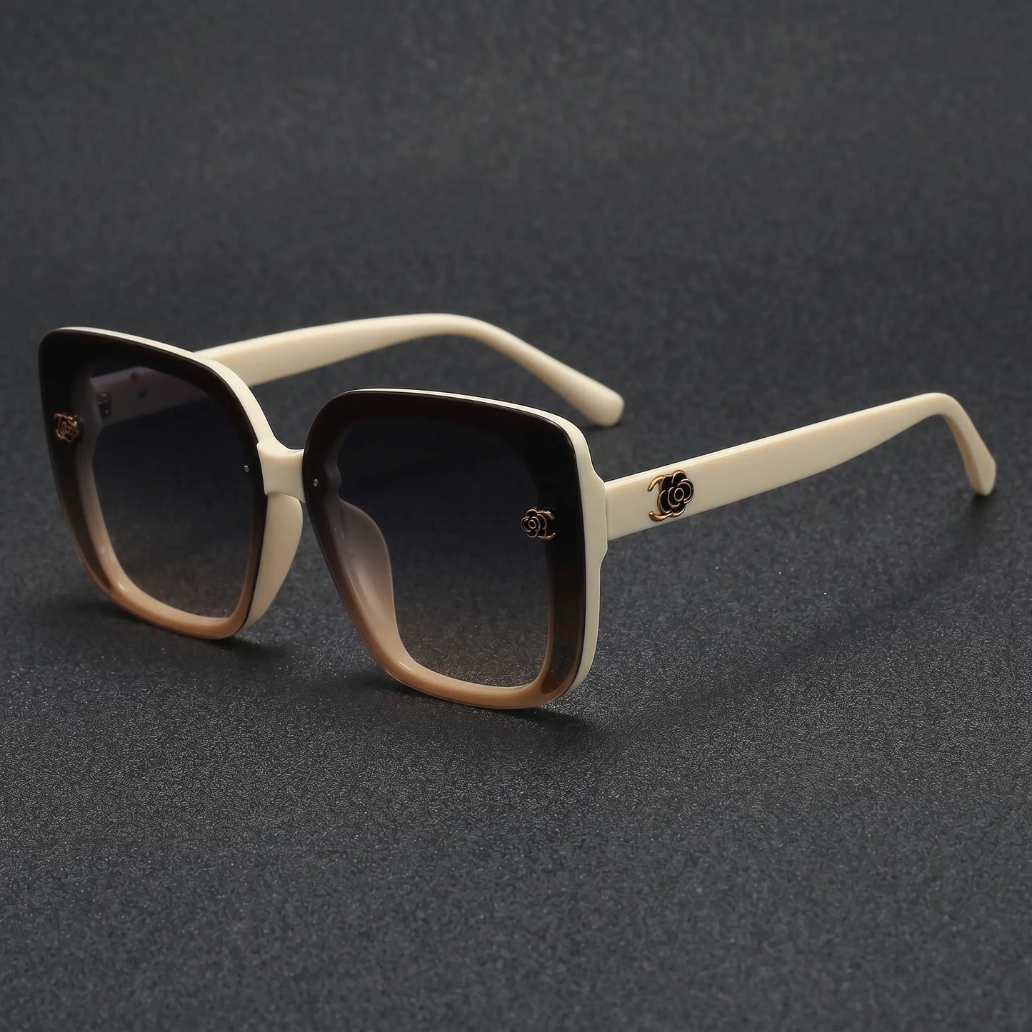Close-up of oversized square sunglasses with off-white temples, featuring a gradient amber-brown front and gradient grey lenses, showcasing a gold floral metal logo detail on the temple.