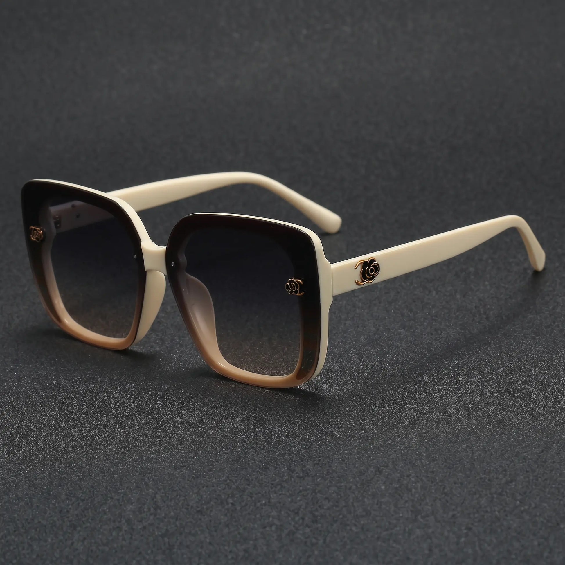 Close-up of oversized square sunglasses with off-white temples, featuring a gradient amber-brown front and gradient grey lenses, showcasing a gold floral metal logo detail on the temple.