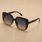 Oversized square sunglasses with a matte black frame, featuring a gradient amber-brown front and dark gradient grey lenses. Embellished with a gold floral-inspired logo on the temples and subtle textured detail on the frame corners.