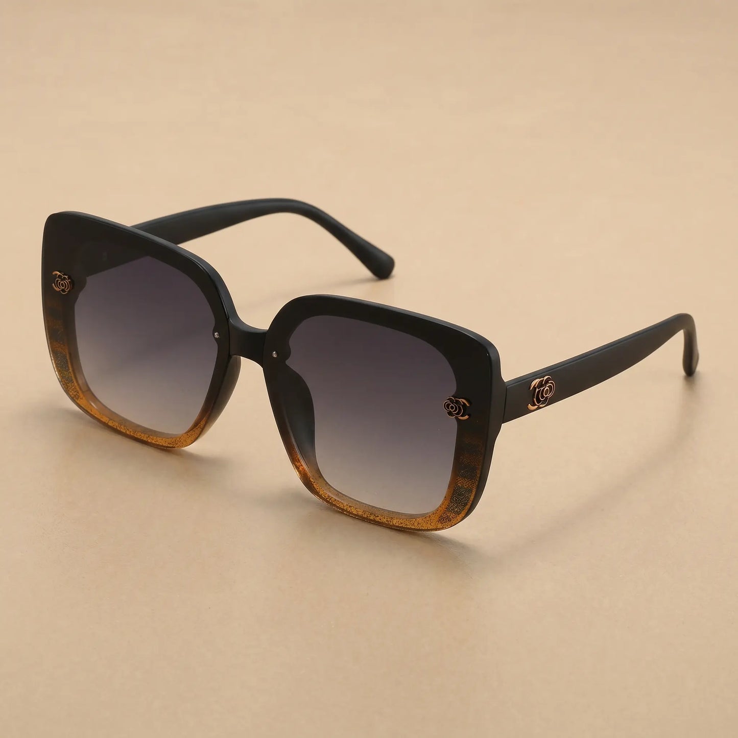 Oversized square sunglasses with a matte black frame, featuring a gradient amber-brown front and dark gradient grey lenses. Embellished with a gold floral-inspired logo on the temples and subtle textured detail on the frame corners.