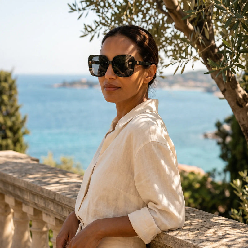 Oversized square Vesryn sunglasses with gradient grey lenses worn by a woman with a serene Mediterranean Sea backdrop, leaning against a stone railing.