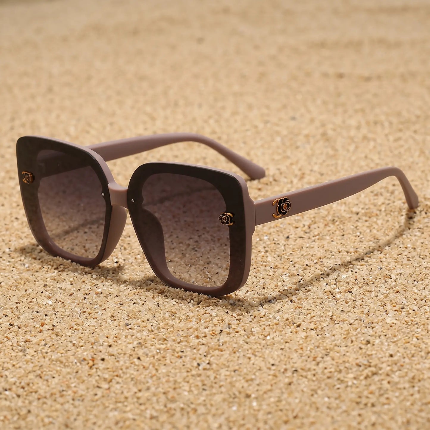 Oversized square sunglasses in a soft taupe frame with black rims and gradient grey lenses, featuring gold floral accents on the temples. A sophisticated fashion accessory.