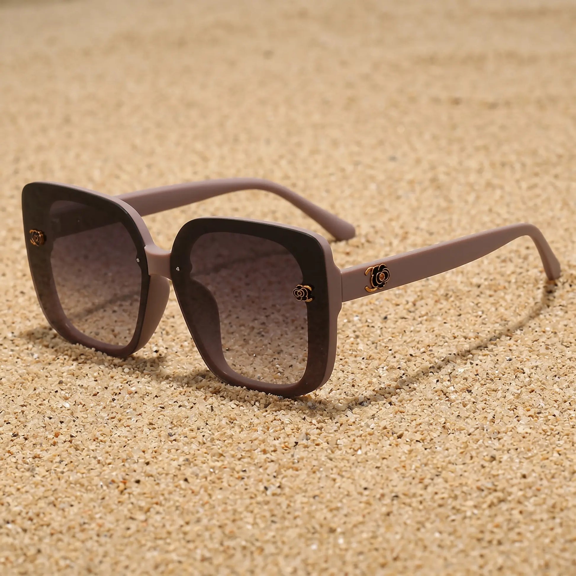 Oversized square sunglasses in a soft taupe frame with black rims and gradient grey lenses, featuring gold floral accents on the temples. A sophisticated fashion accessory.
