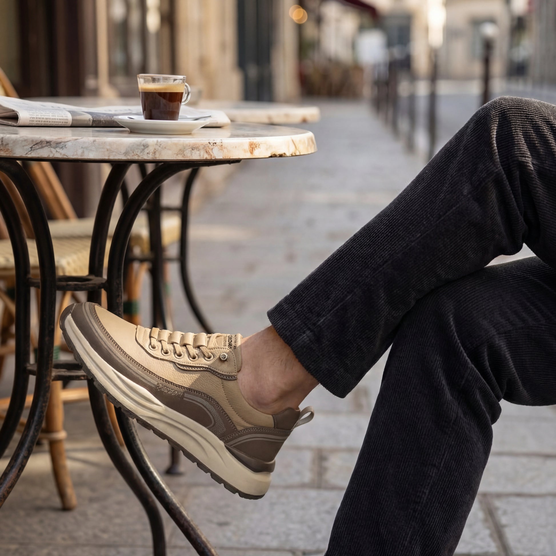 Vexor shock-absorbing urban sneakers in khaki worn by a man sitting at a marble cafe table with espresso, showing the thick EVA sole profile.