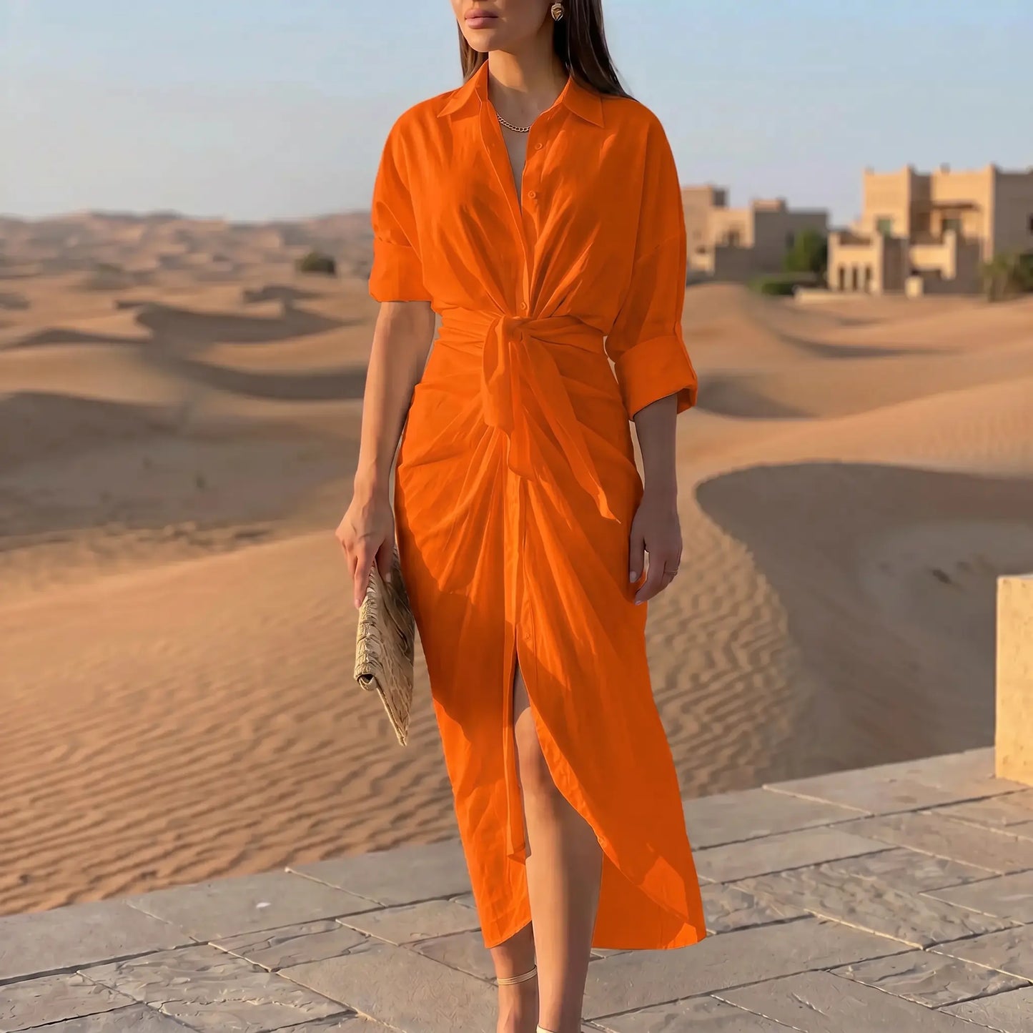 Model wearing a vibrant orange tie-waist midi shirt dress with a pointed collar, rolled sleeves, and a draped tulip skirt, styled in a desert architectural setting.