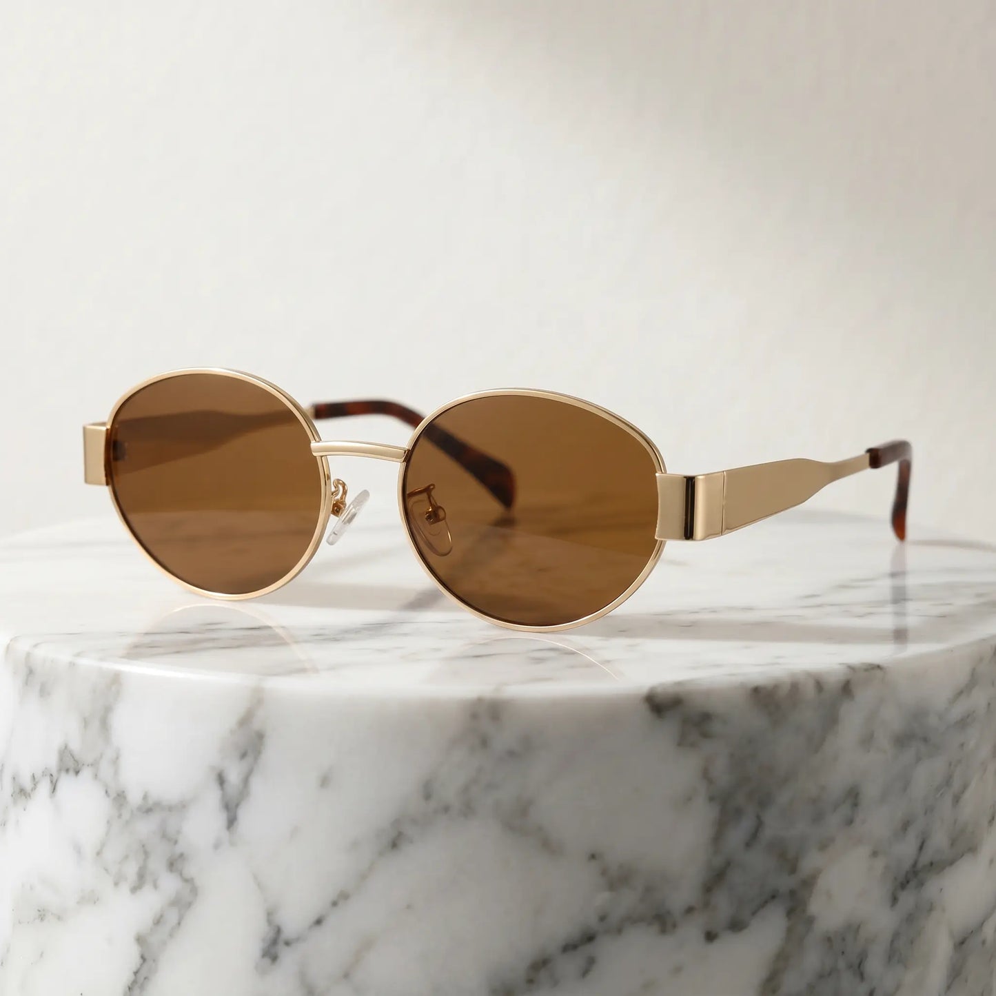 Gold-toned vintage oval metal frame sunglasses with brown tinted lenses and tortoiseshell temple tips, presented on a marble pedestal.