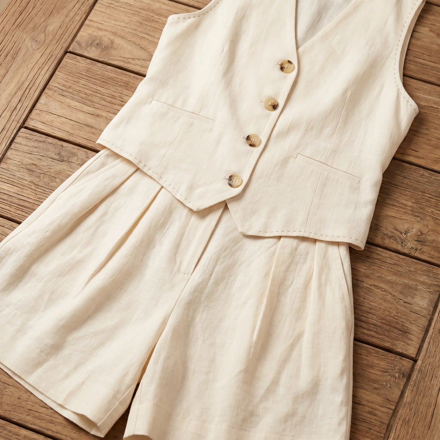A creamy ecru linen-blend vest and wide-leg shorts set displayed flat on a wooden surface, showcasing the V-neck vest with mother-of-pearl buttons and pleated high-waisted shorts.