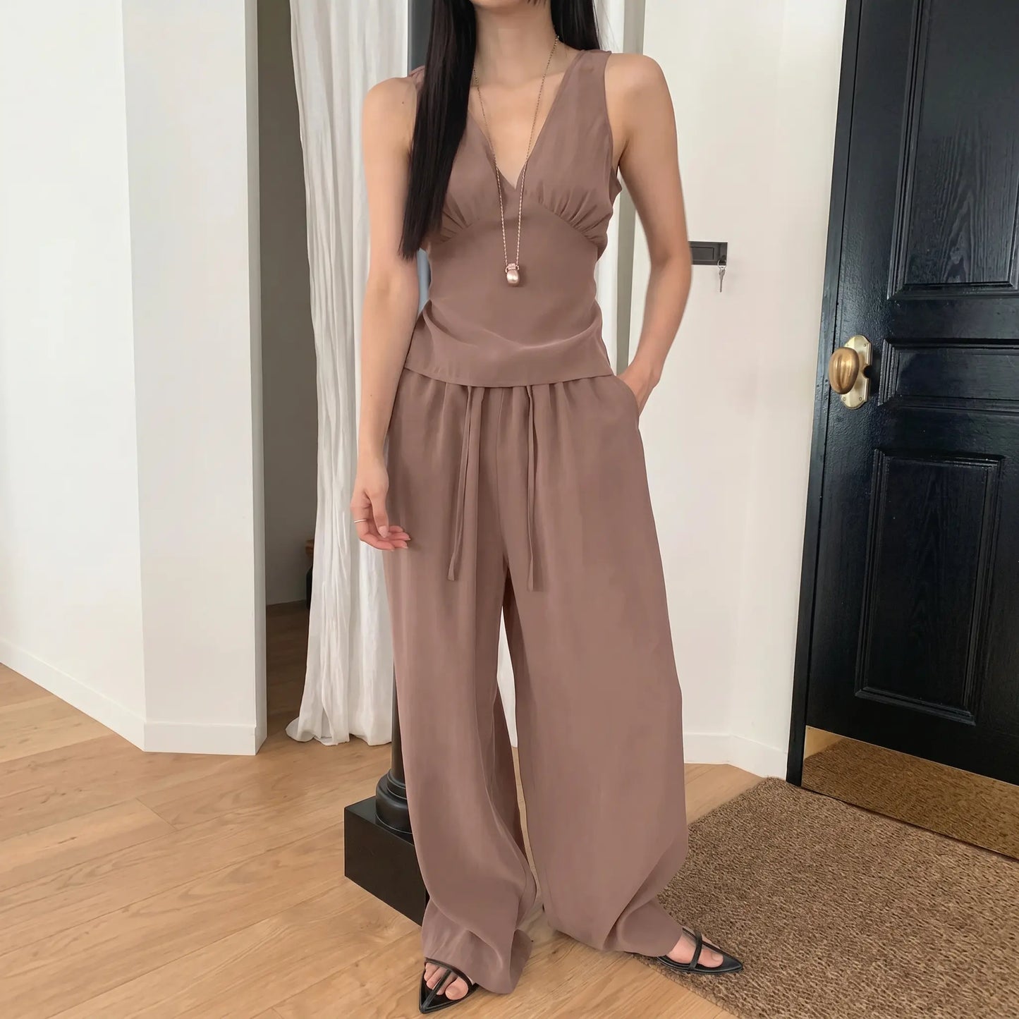 A woman models a two-piece taupe resort set featuring a sleeveless V-neck top with gathered bust and flowy wide-leg trousers with an elastic drawstring waist.