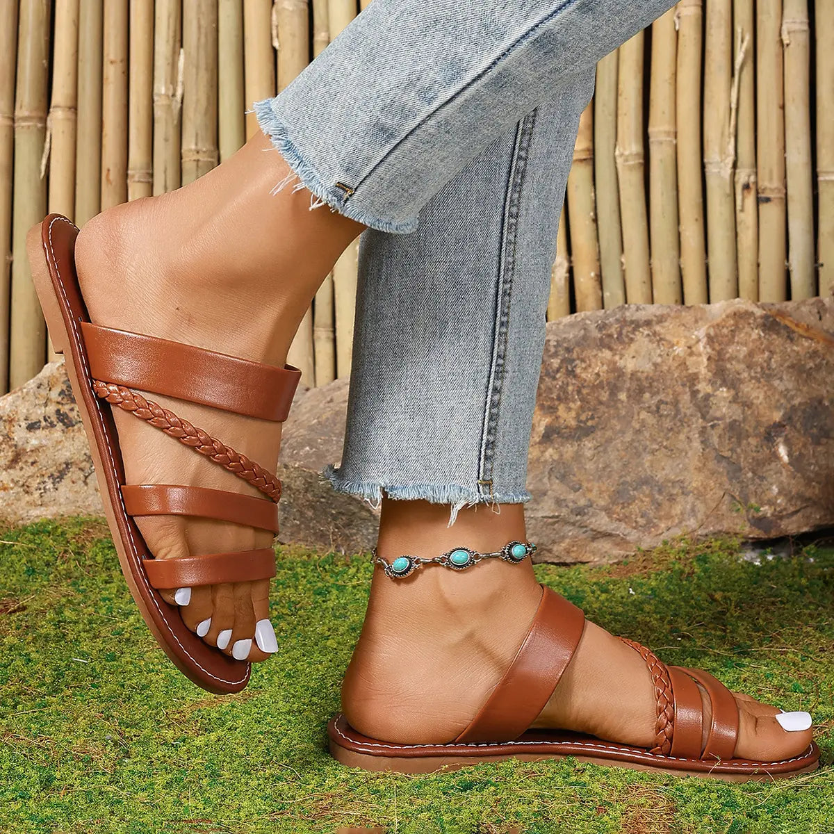 Vorlag women's brown braided strap summer slide sandals worn with distressed denim jeans and a turquoise anklet in a natural outdoor setting.