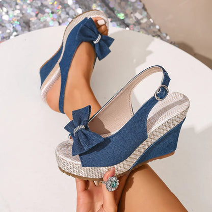 Vykos denim-look wedge espadrille sandals featuring a sparkling bow detail and adjustable slingback strap, shown on a foot with a second sandal held by a hand.