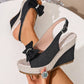 Close-up view of a Vykos black denim-look wedge sandal featuring a crystal-embellished bow, espadrille jute-wrapped wedge and platform, and an adjustable slingback strap with a metal buckle.