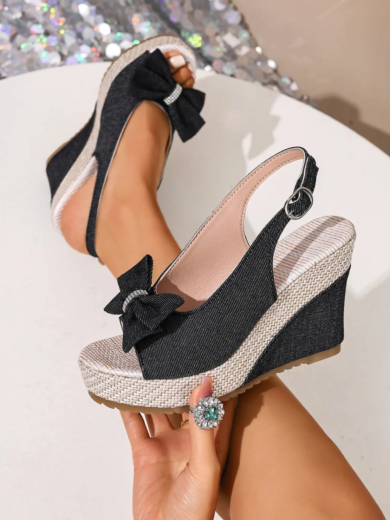 Close-up view of a Vykos black denim-look wedge sandal featuring a crystal-embellished bow, espadrille jute-wrapped wedge and platform, and an adjustable slingback strap with a metal buckle.