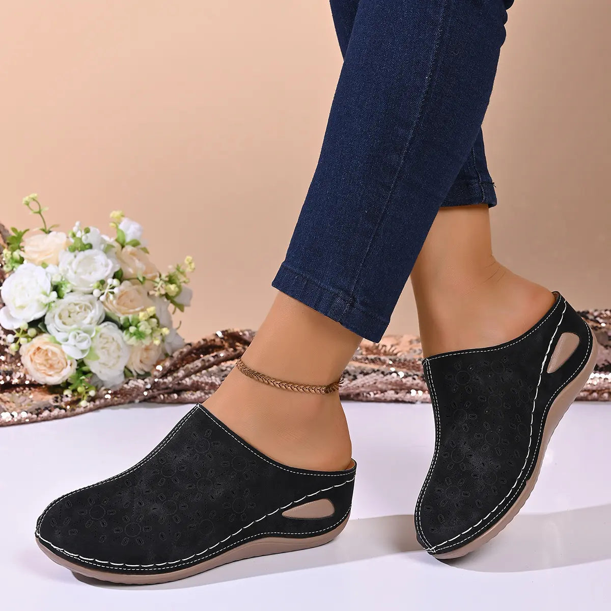 Black perforated wedge mules worn by a model, showcasing the comfortable slip-on design with a rounded toe and medium wedge heel, styled with denim jeans.