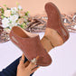 Close-up of women's Vylos perforated wedge mules in a warm brown hue, shown on a model's foot with intricate floral laser-cut details and contrasting white stitching. A hand holding the shoe displays a decorative floral ring.