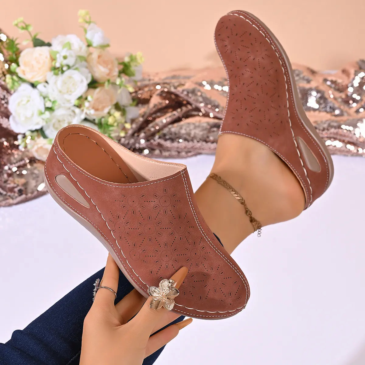 Close-up of women's Vylos perforated wedge mules in a warm brown hue, shown on a model's foot with intricate floral laser-cut details and contrasting white stitching. A hand holding the shoe displays a decorative floral ring.