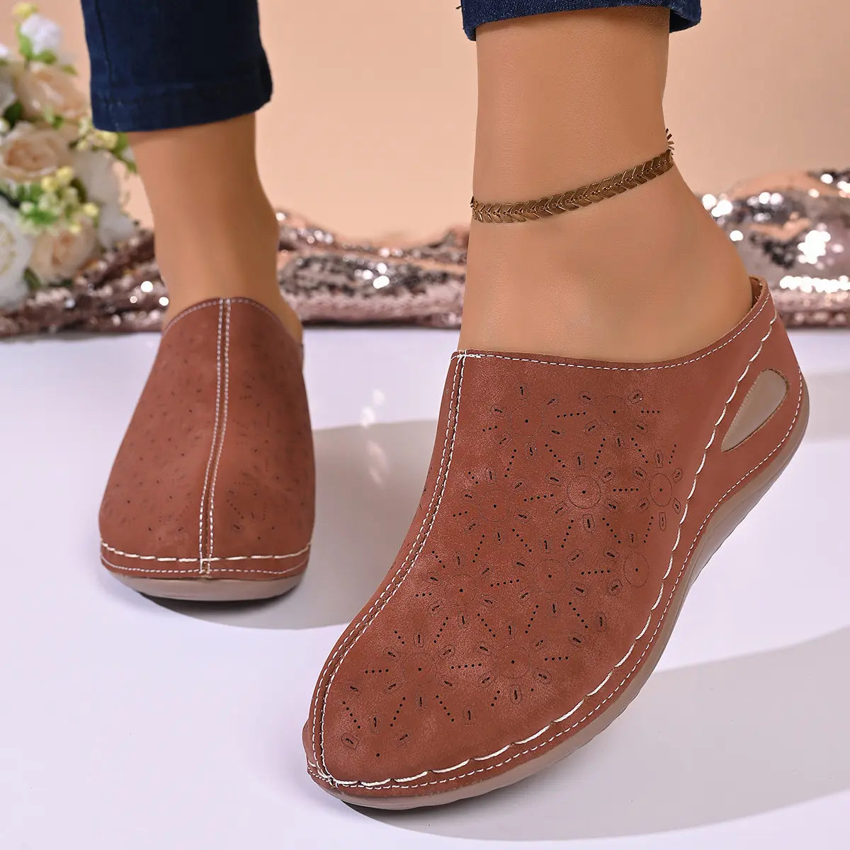 Vylos women's perforated floral wedge mules in rich earth brown, shown on a model's feet from a three-quarter front view, highlighting the intricate laser-cut details and white stitching.
