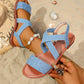 Blue textured Roman flat sandals with criss-cross elasticated straps and a polished silver ring embellishment, styled on a foot with an anklet on a sandy beach. Features a fish mouth peep-toe and flat sole for summer wear.