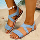 Vytka light blue textured fabric Roman flat sandals with elasticated criss-cross straps and a polished silver-tone ring embellishment, shown in a beach lifestyle setting.