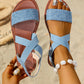 Close-up of light blue textured Roman flat sandals with elasticated criss-cross straps and a polished silver ring embellishment on a sandy beach background, showcasing a model's feet adorned with jewelry.