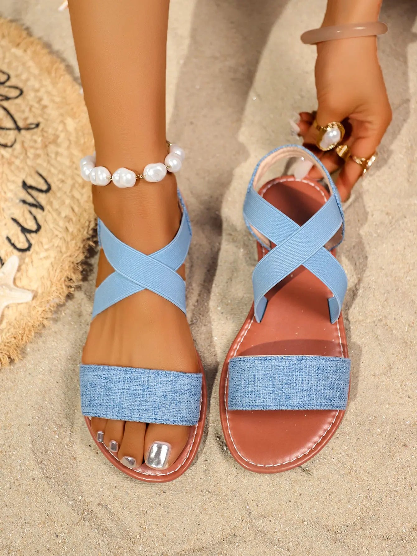 Vytka Roman flat sandals in light blue textured fabric with criss-cross elasticated slingback straps and a silver-tone metal ring embellishment, styled on sand with beach accessories.