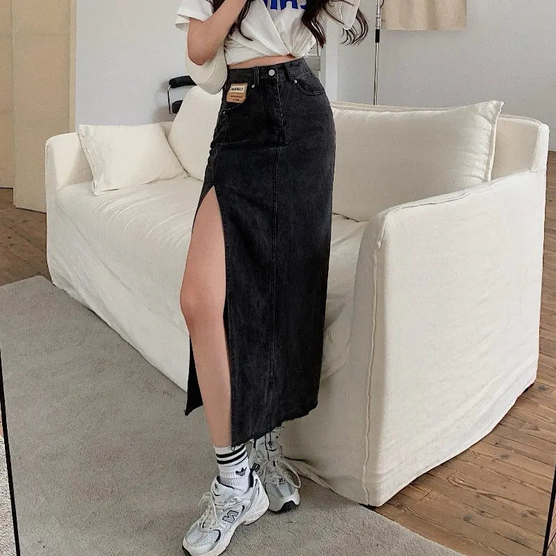 Zubari washed obsidian high-waisted denim maxi skirt with a dramatic front slit and raw frayed hem. Worn by a model in a casual indoor setting, showcasing the tan patch detail.