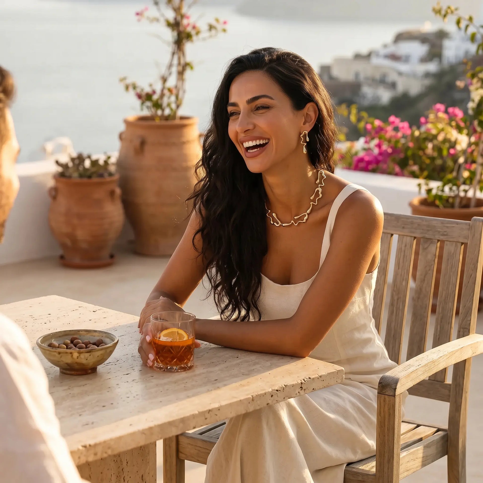 Smiling woman wearing an exquisite Wexari gold-toned sculptural liquid metal collar necklace and matching abstract teardrop earrings on an elegant outdoor terrace.