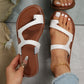 Close-up lifestyle view of elegant white asymmetrical strap flat slide sandals worn on a foot, featuring a brown footbed, white contrast stitching on the sole, and a gold anklet.
