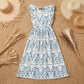 Bohemian white midi dress with blue floral print, featuring ruffled cap sleeves, a tie-back open back, and an elasticated waist, styled with straw hats on a jute rug.
