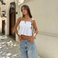 Woman wearing a white broderie anglaise camisole top with a ruffled neckline and tie straps, styled with high-waisted jeans in an outdoor urban setting.