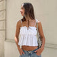 A young woman in a white broderie anglaise camisole top with ruffled neckline and tie straps, styled with denim jeans and a shoulder bag. The top features a babydoll silhouette and delicate floral eyelet embroidery.