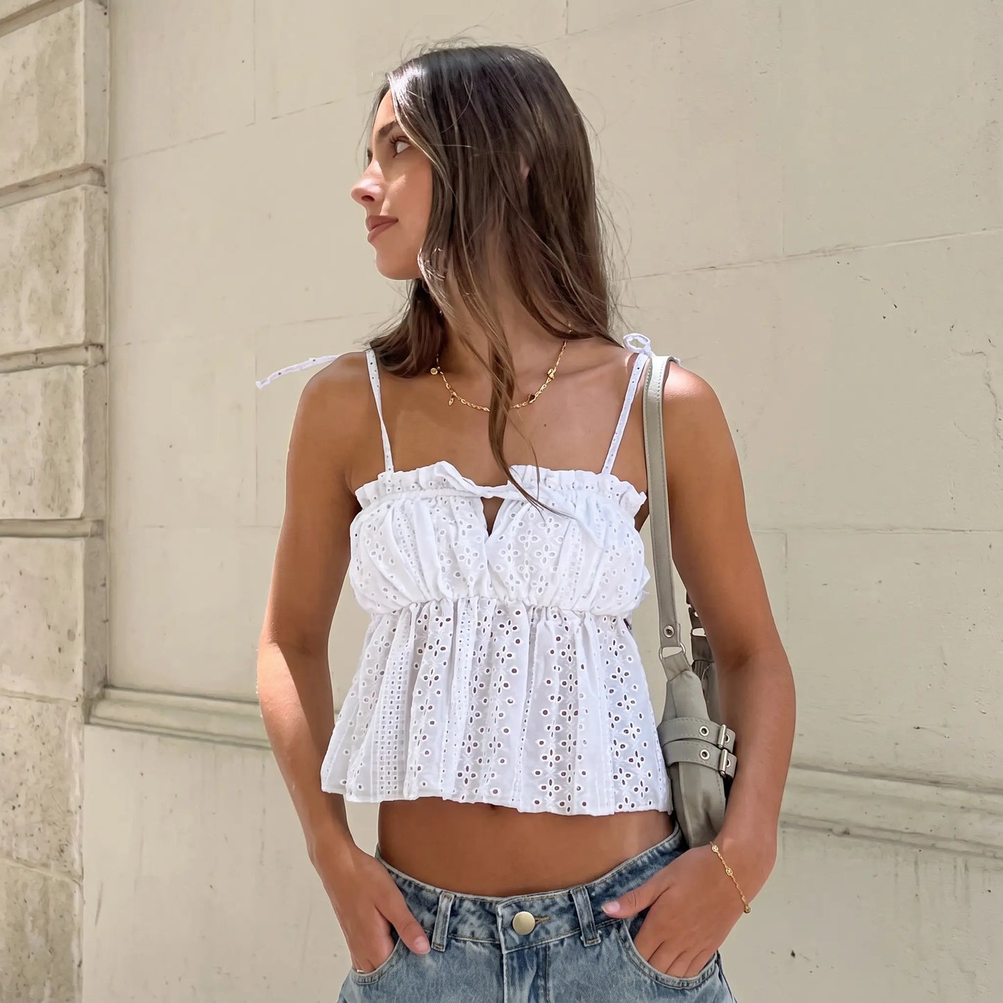 A young woman in a white broderie anglaise camisole top with ruffled neckline and tie straps, styled with denim jeans and a shoulder bag. The top features a babydoll silhouette and delicate floral eyelet embroidery.