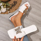 White flat slide sandals featuring a gold-tone butterfly embellishment with crystal accents, worn by a model on feet with painted toenails, styled with a straw hat and flowers on a wooden floor.
