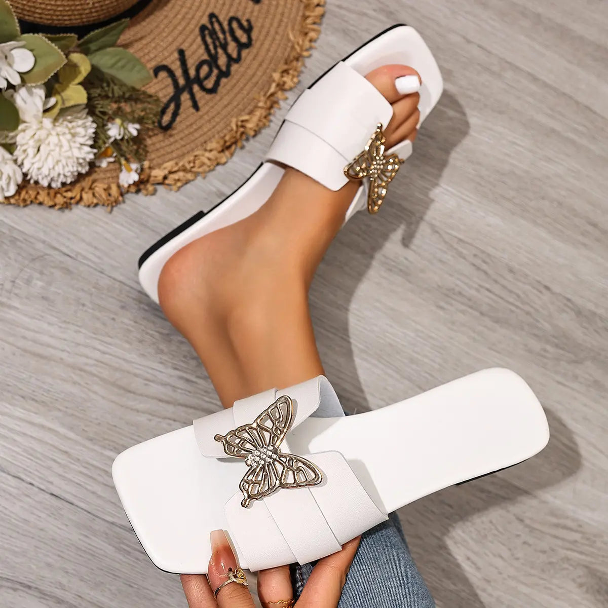 White flat slide sandals featuring a gold-tone butterfly embellishment with crystal accents, worn by a model on feet with painted toenails, styled with a straw hat and flowers on a wooden floor.