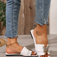 White Aethel flat slide sandals adorned with a gold butterfly embellishment, worn by a model with jeans and a gold anklet on a wooden floor.