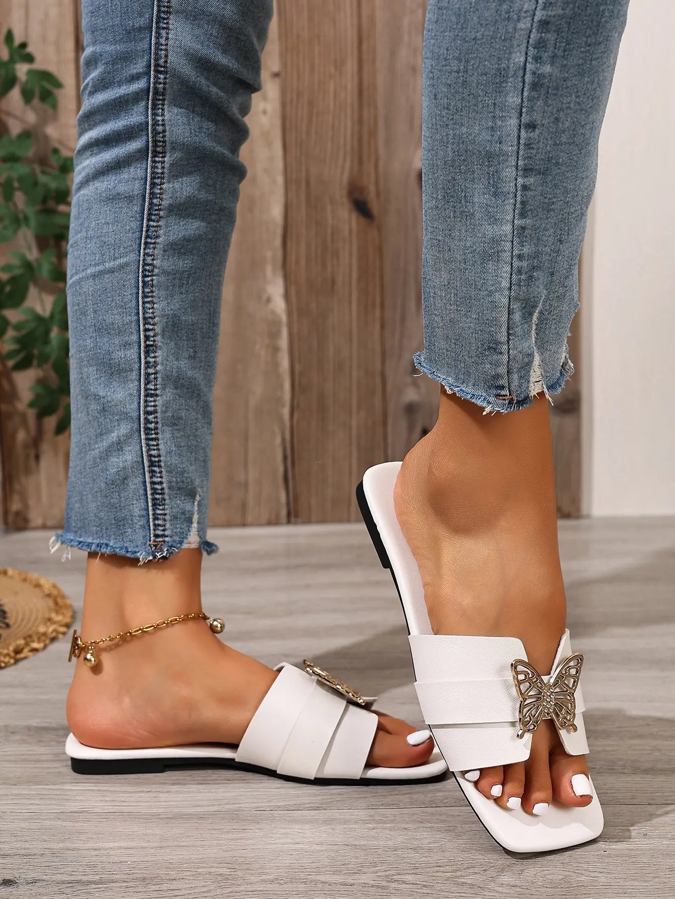 White Aethel flat slide sandals adorned with a gold butterfly embellishment, worn by a model with jeans and a gold anklet on a wooden floor.