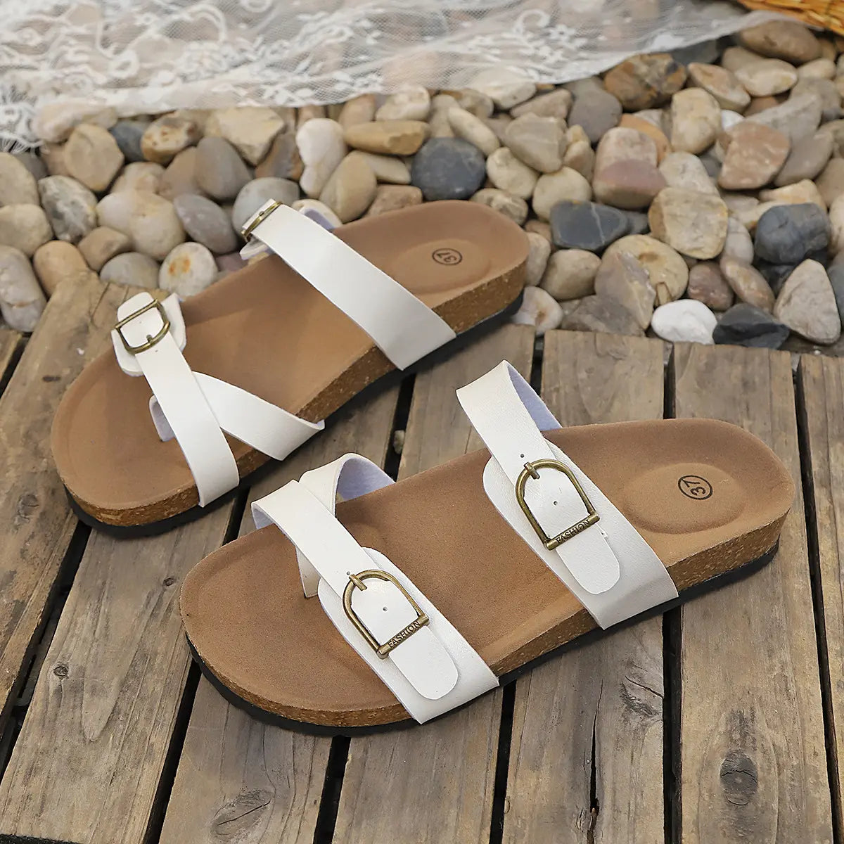 Pair of white Lyraea cork footbed slide sandals with adjustable antique brass buckle straps, shown flat on a rustic wooden surface with stones and lace.